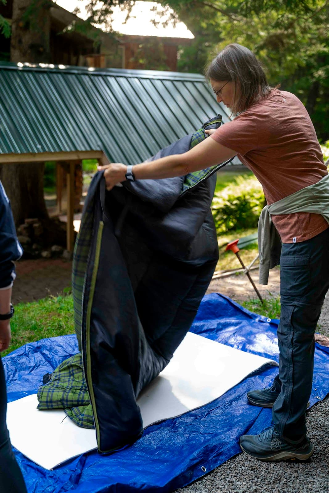 A camper unfolding a sleeping bag above a foam sleeping mat on a blue groundsheet.
